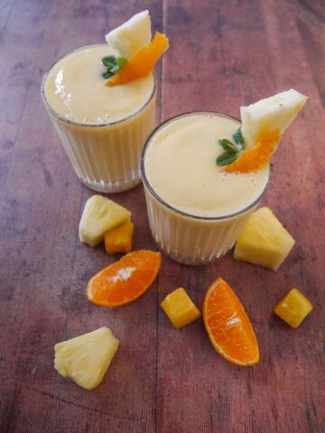 Two glasses of tropical mango pineapple smoothie garnished with a wedge of fresh pineapple and a slice of clementine.
