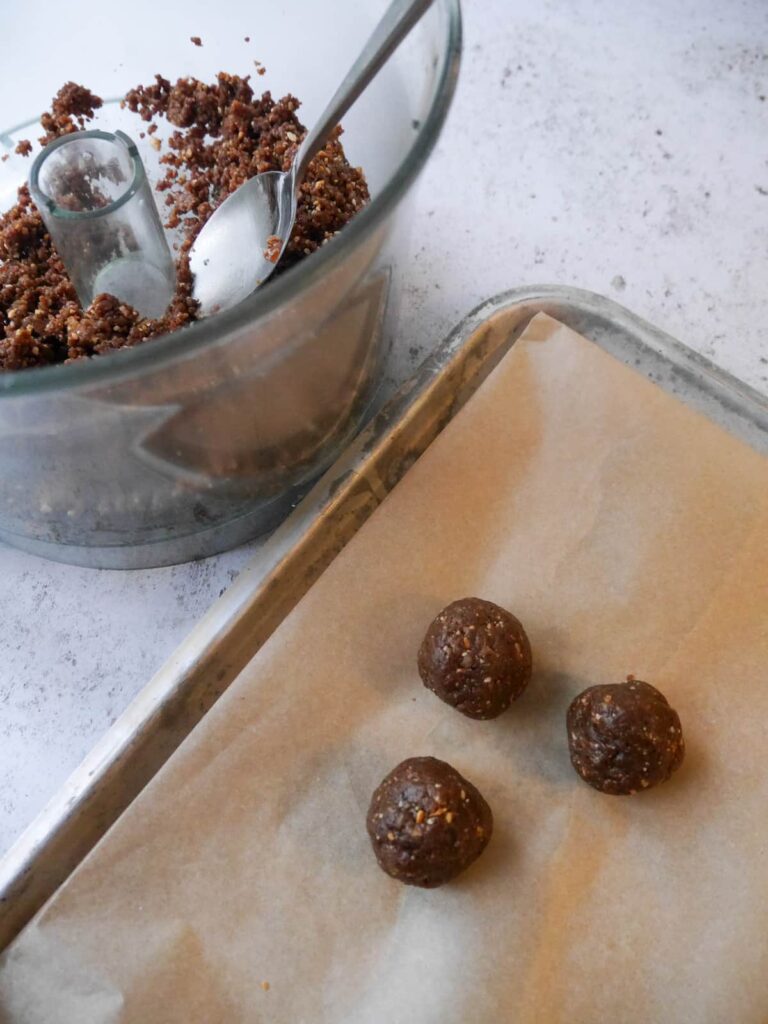 A paper lined tray with date energy balls, with food process bowl of mixture set alongside.
