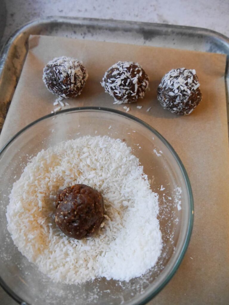 A paper lined tray with coconut coated energy balls with a glass bowl of desiccated coconut set on top.