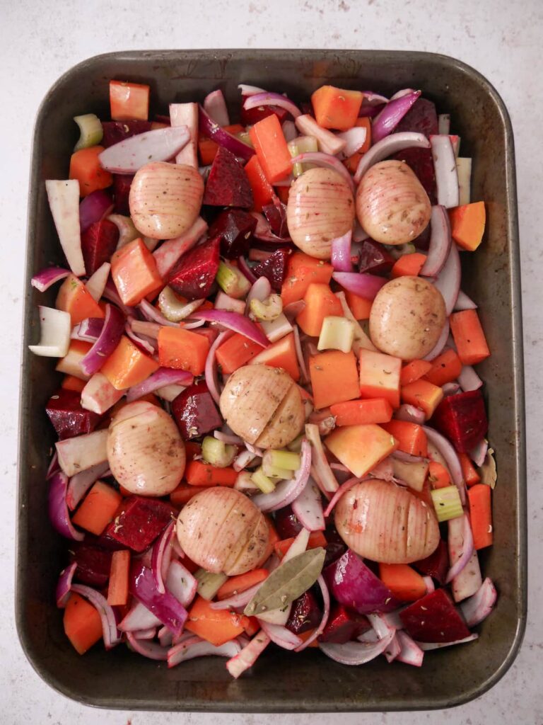 A large oven tray filled with chopped root vegetables, onion, garlic, potatoes and with added bay leaves and thyme.