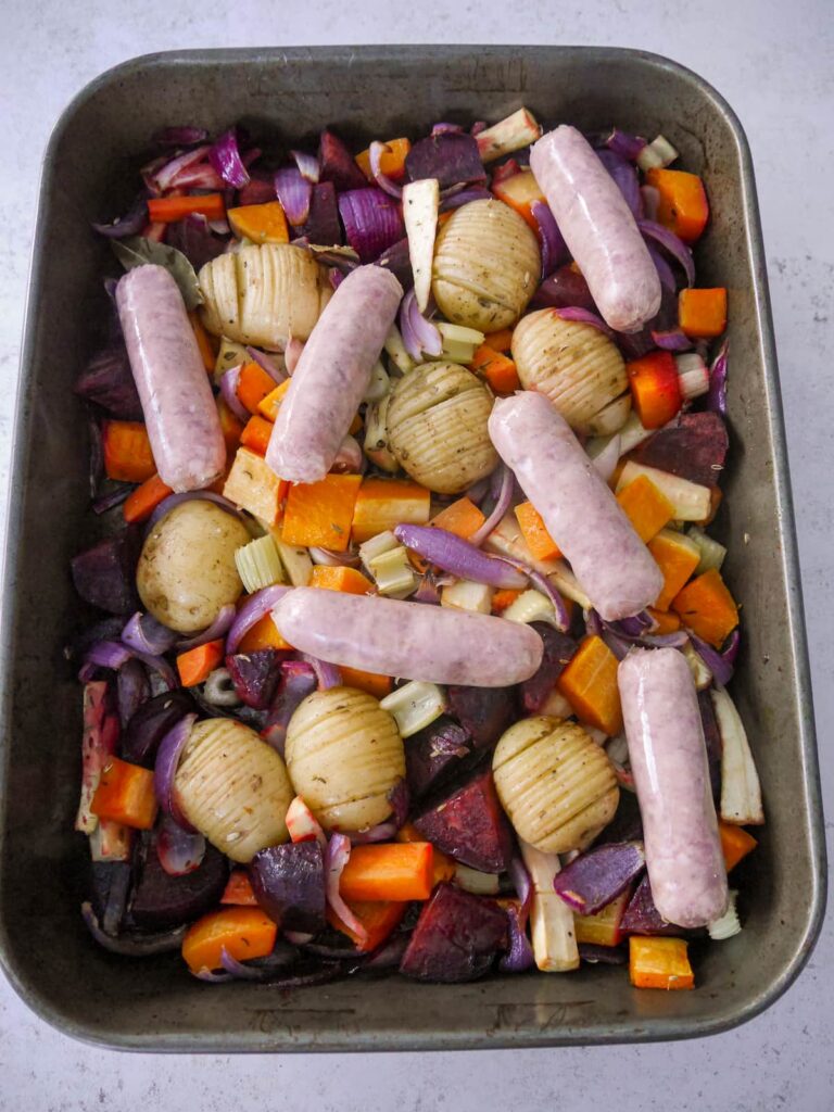A tray of part roasted vegetables and potatoes, topped with pork sausages.