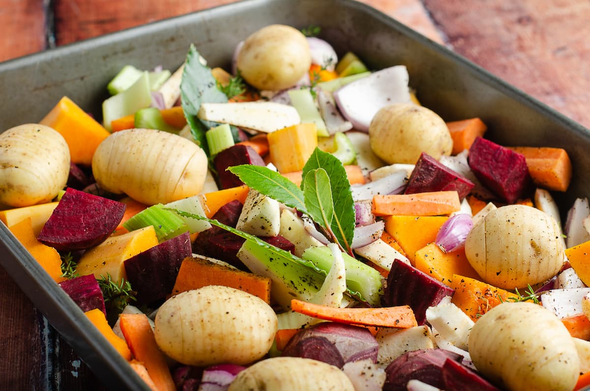 A large oven proof dish filled with raw root vegetables, hassleback potatoes and fresh herbs.