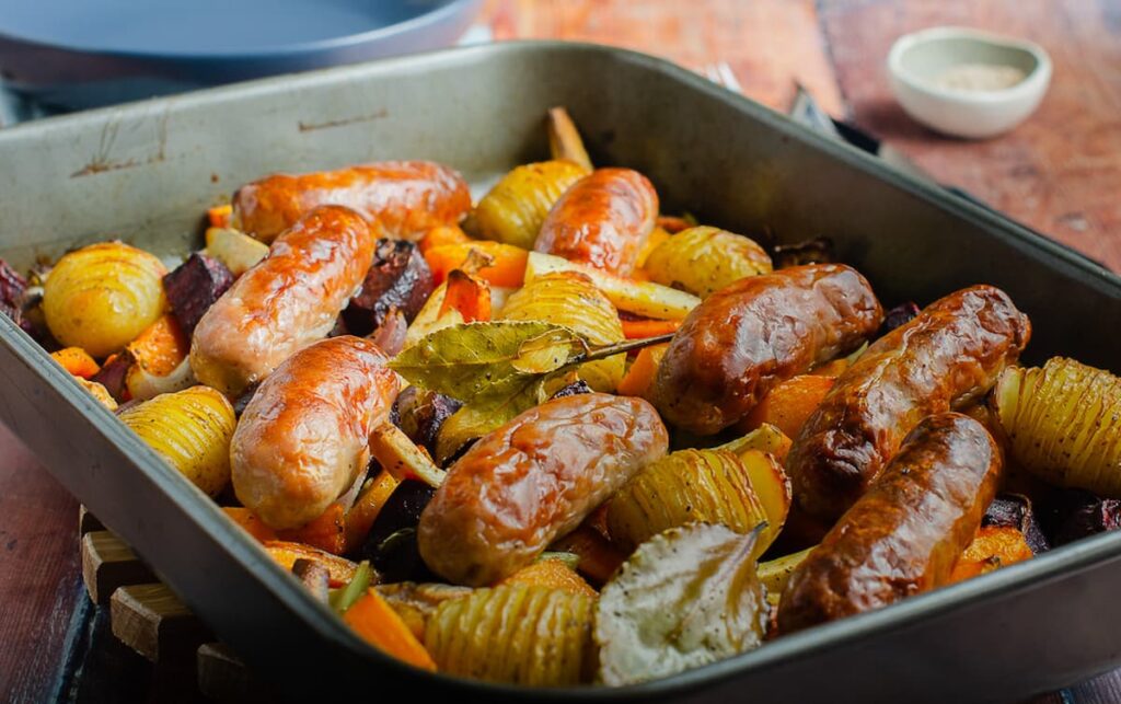 A large oven proof dish filled with roasted vegetables, hassleback potatoes and cooked sausages.