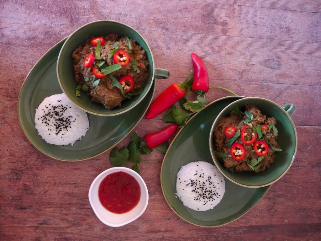 Two green bowls filled with beef rendang garnishes with sliced red chilli and chopped coriander leaf, with a portion of steamed white rice, garnished with nigella seeds and a bowl of sambal set alongside.