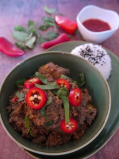 A green bowl filled with beef rendang garnishes with sliced red chilli and chopped coriander leaf, with a portion of steamed white rice, garnished with nigella seeds and a bowl of sambal set alongside.