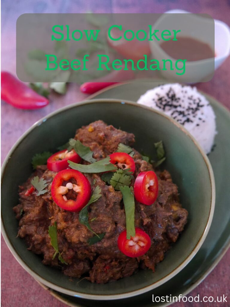 Pinnable image with recipe title and a green bowl filled with beef rendang garnishes with sliced red chilli and chopped coriander leaf, with a portion of steamed white rice, garnished with nigella seeds set alongside.