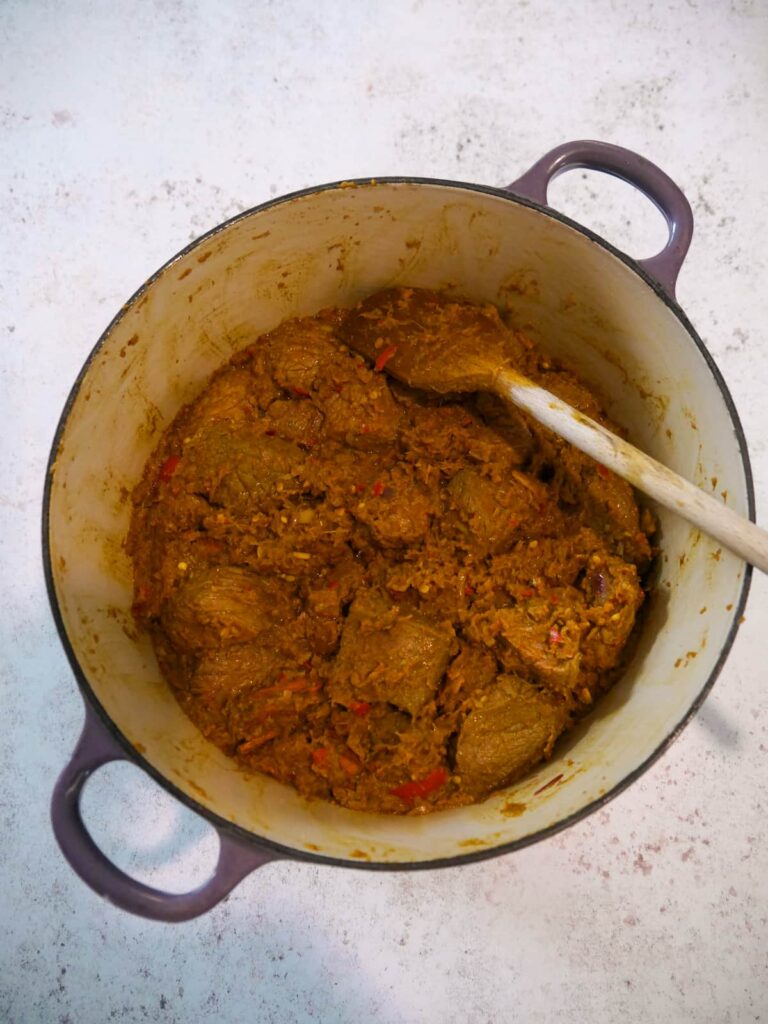 Dutch oven with vegetable and spice base ingredients with added chunks of beef with the paste mixture coating the beef.