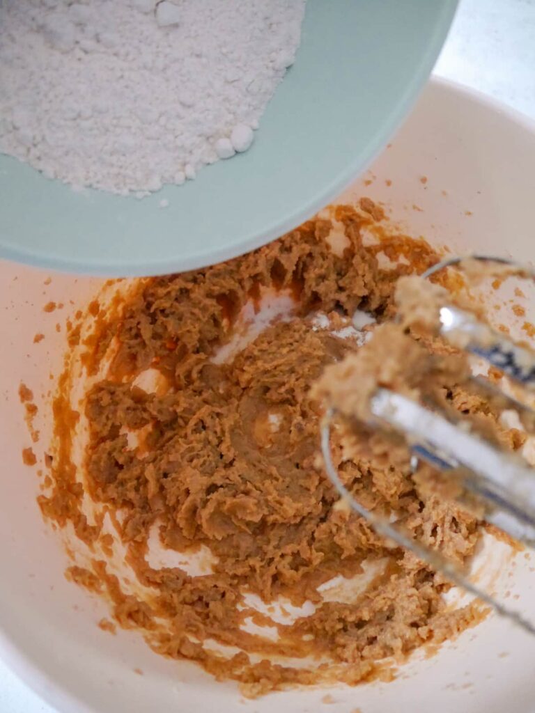 A white bowl of creamed sugar, butter and egg with flour being added to whisk in.