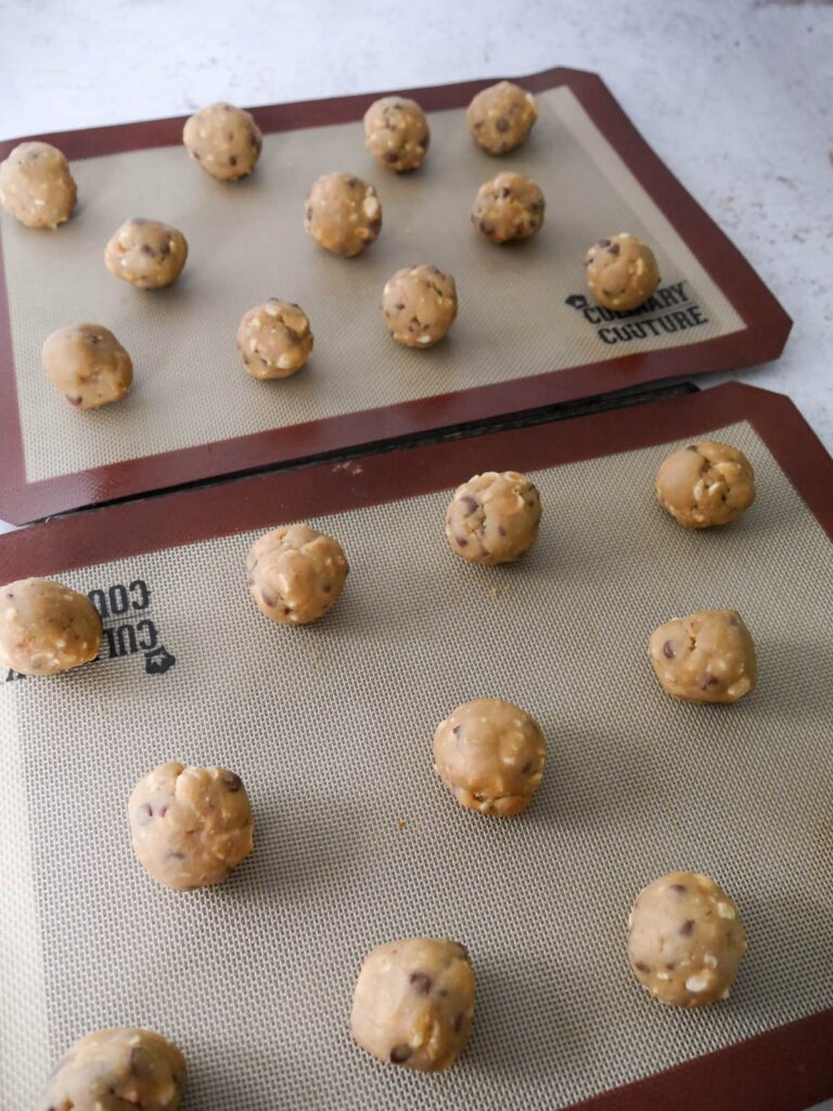 Two cookie sheets lined with silicone liners with balls of cookie dough laid on top.