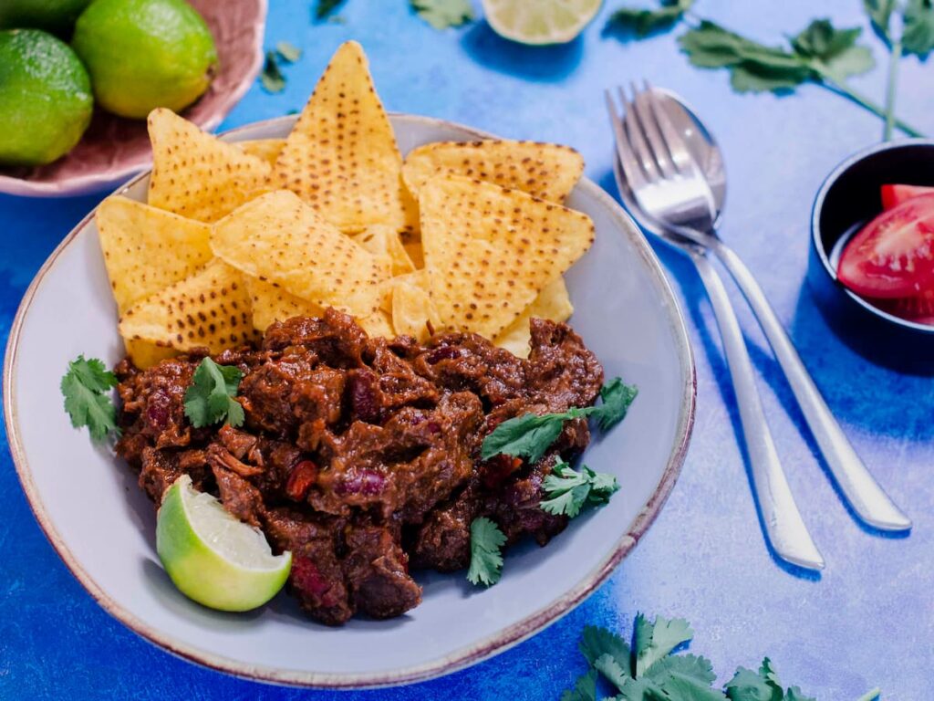 A bowl of slow cooked chunky beef chilli served with tortilla chips and a wedge of lime.