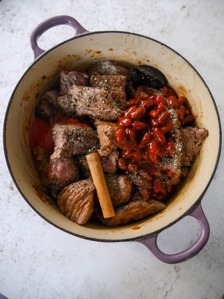 A Dutch oven filled with sauteed vegetables with added seared beef chunks, kidney beans in a chilli sauce and cinnamon stick.