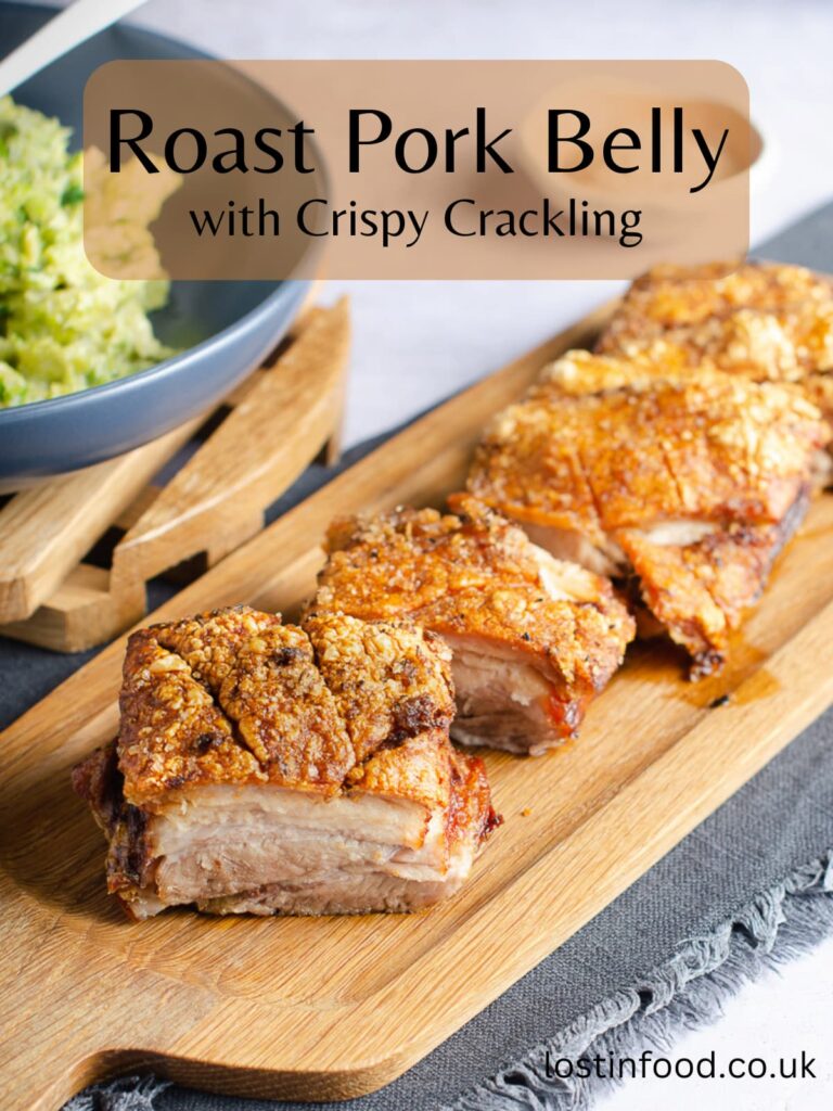 Pinnable image with recipe title and image of portions of roasted pork belly with crispy crackling top set out on a wooden board.