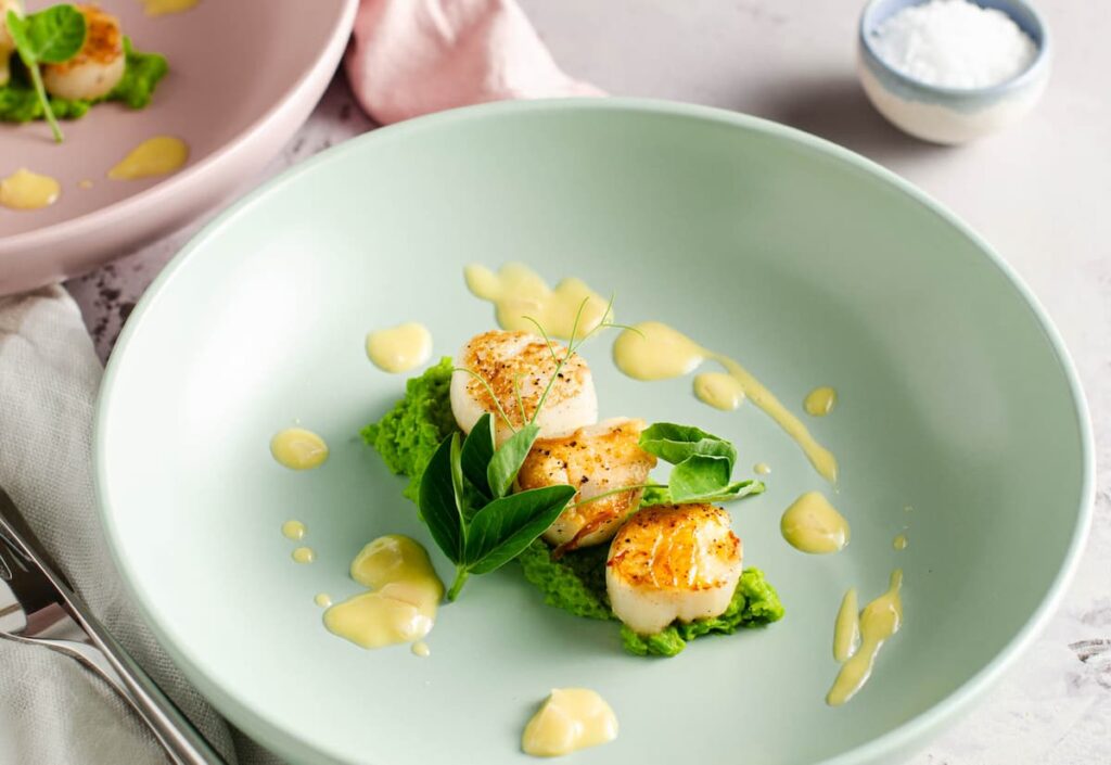 A blue bowl with pea puree, topped with three seared scallops with a drizzle of butter sauce and garnished with fresh pea shoots.
