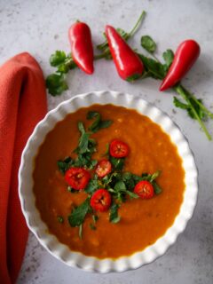 A white bowl of spicy red lentil soup garnished with chopped coriander leaf and thinly spiced red chilli.