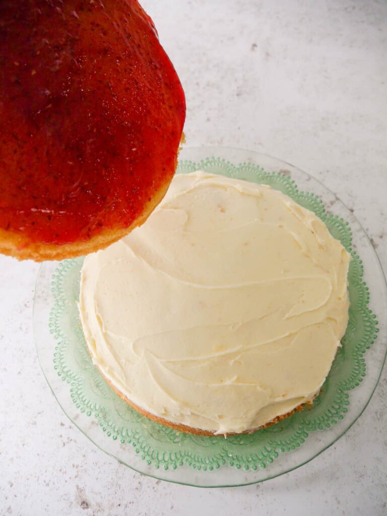 A sponge cake topped with a layer of butter icing with a second sponge cake spread with raspberry jam being laid on top.