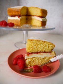 A slice of Victoria sponge on a pink plate with added fresh raspberries and the whole cake on a cake stand in the background.