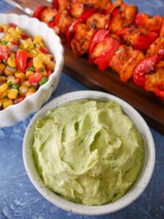 Bowl of avocado cream with a bowl of corn salsa and wooden board with lemon paprika chicken kebabs set alongside.