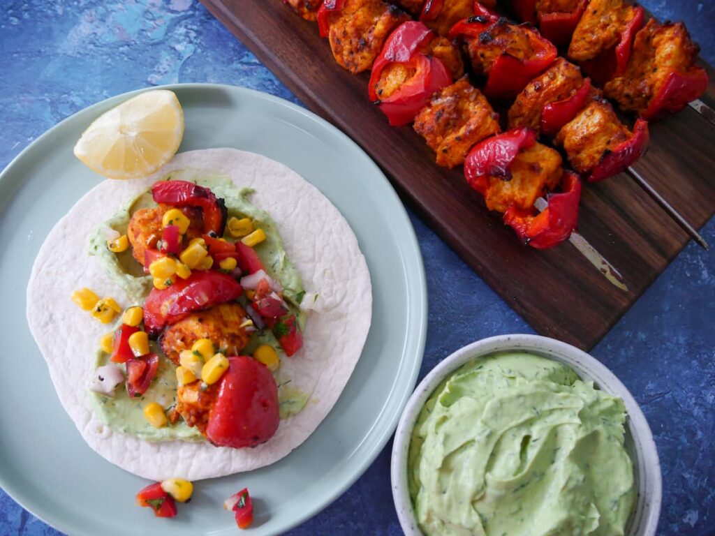 A plate with chicken taco served with avocado cream and topped with corn salsa, with a board of lemon paprika chicken kebabs and a bowl of avocado cream set alongside.