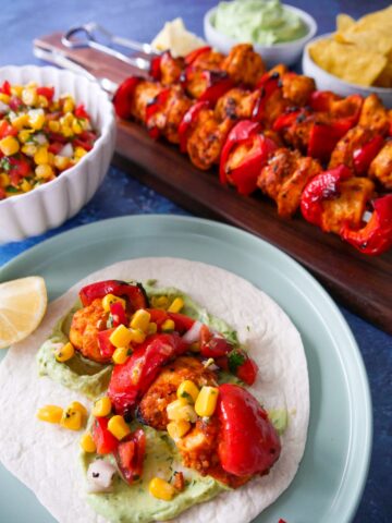 A plate with chicken taco served with avocado cream and topped with corn salsa, with a board of lemon paprika chicken kebabs and a bowl of corn salsa set alongside.