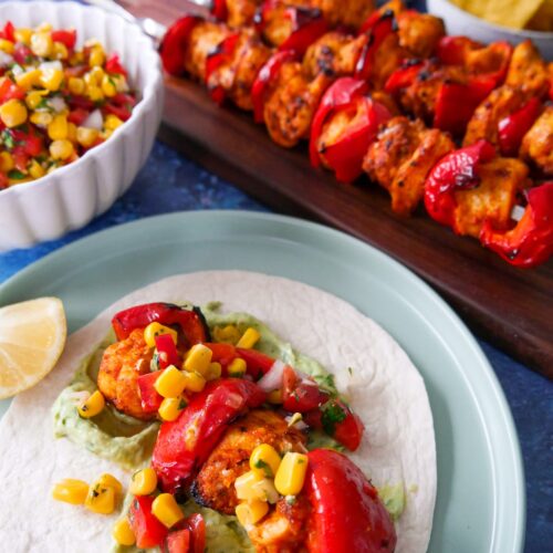 A plate with chicken taco served with avocado cream and topped with corn salsa, with a board of lemon paprika chicken kebabs and a bowl of corn salsa set alongside.