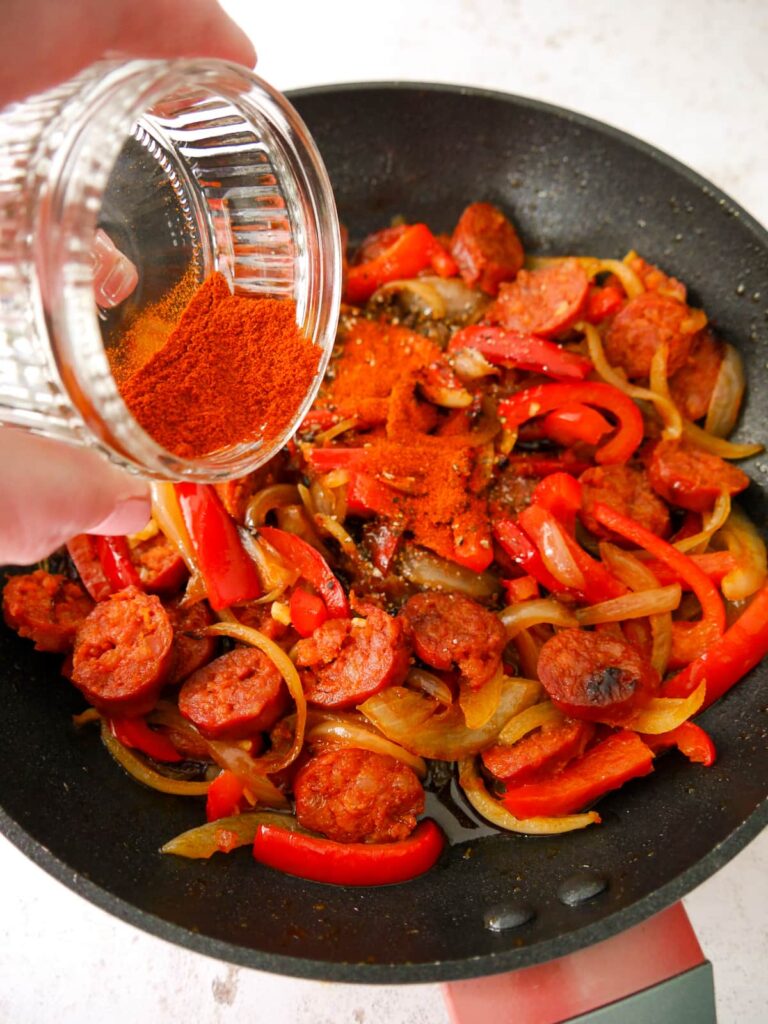 A non stick skillet with cooked onions, red pepper, garlic and sliced chorizo sausage with smoked paprika being added.