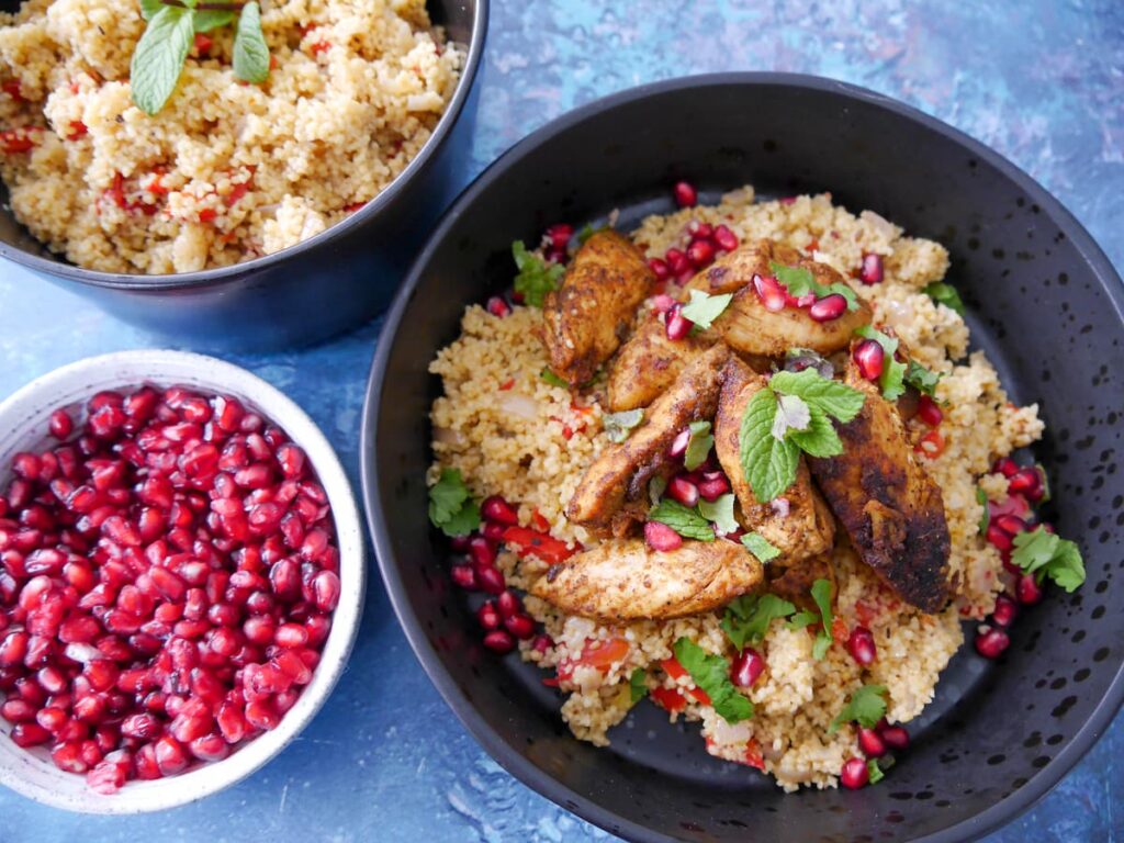 A black bowl filled with spicy couscous topped with ras-el-hanout chicken and garnished fresh herbs and pomegranate seeds, with a bowl of spicy couscous and bowl of pomegranate seeds set alongside.