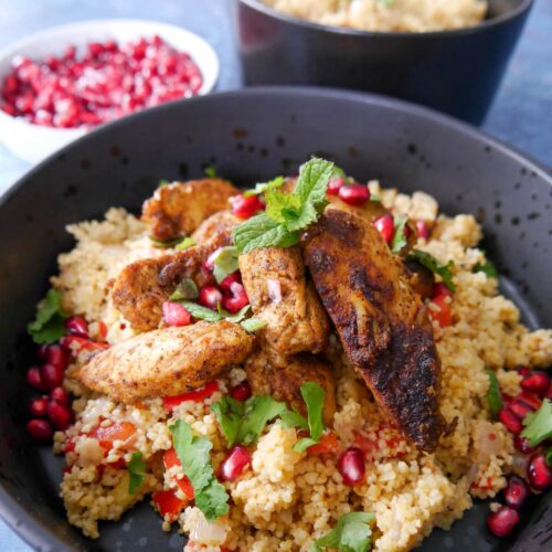 A black bowl filled with spicy couscous topped with ras-el-hanout chicken and garnished fresh herbs and pomegranate seeds, with a bowl of pomegranate seeds set alongside.