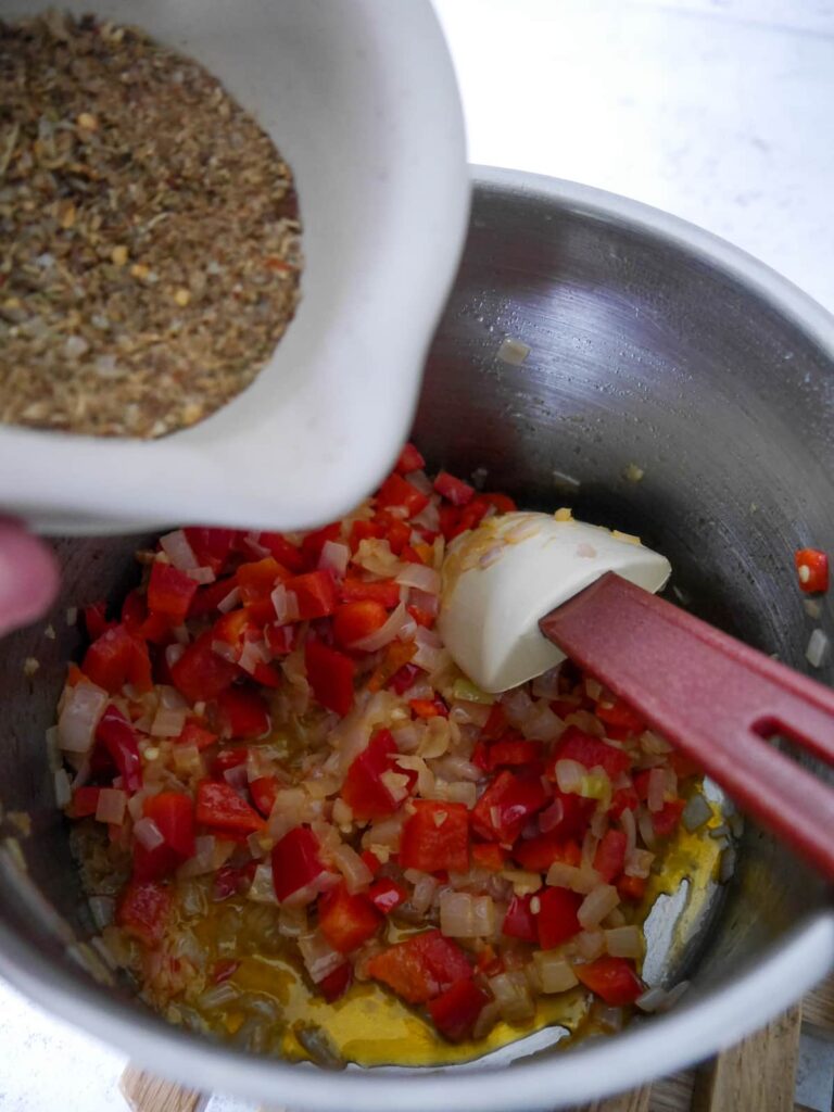 A pan of sauteed shallot, red bell pepper, chilli and garlic with dry crushed spices being added.
