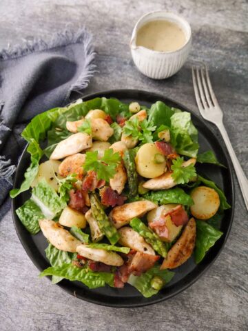 A black plate topped with a salad of lettuce leaves, sauteed chicken, asparagus, new potatoes and bacon with a small just of salad dressing set alongside.