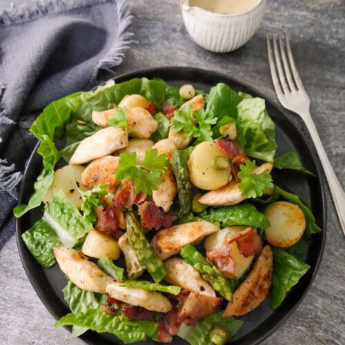 A black plate topped with a salad of lettuce leaves, sauteed chicken, asparagus, new potatoes and bacon with a small just of salad dressing set alongside.