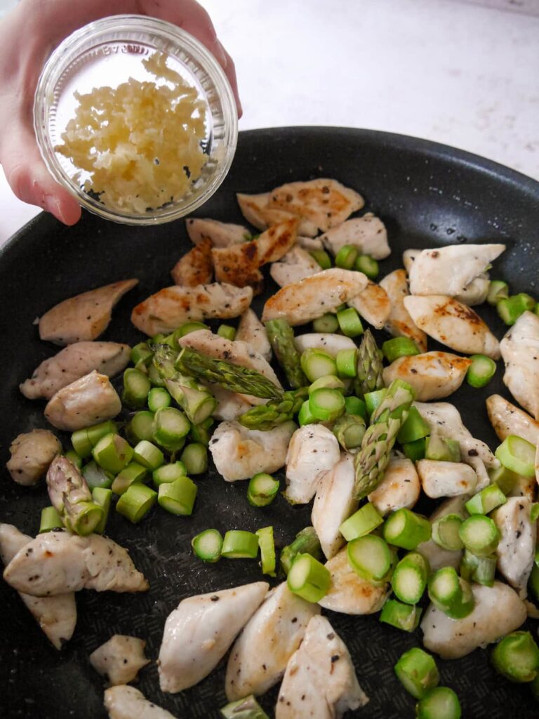 A frying pan with sauteed chicken strips, with asparagus and crushed garlic being added.