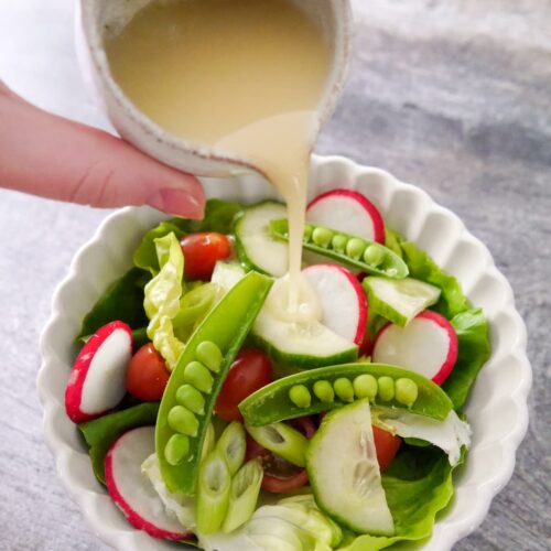 A small jug of French dressing being poured over a white bowl filled with salad of lettuce, radish, tomato, cucumber, spring onions and sugar snap peas.