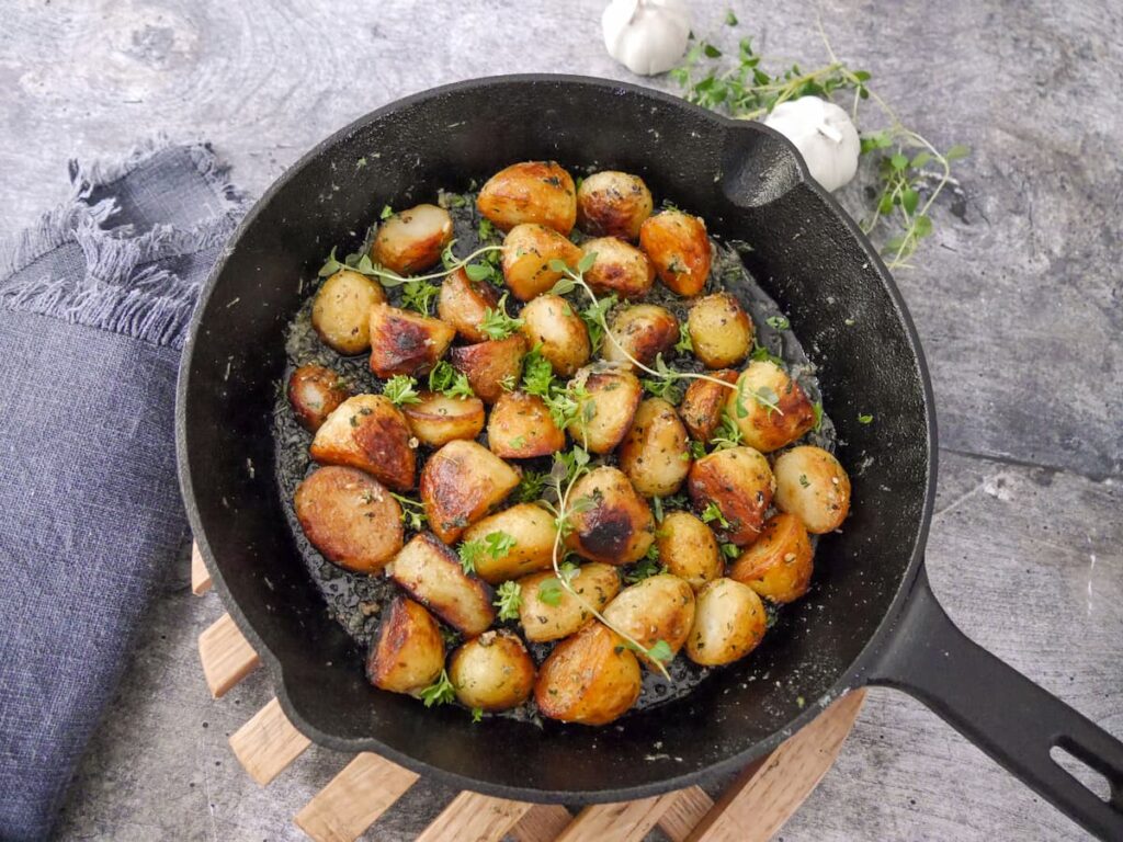 A cast iron skillet set on a wooden trivet, filled with fried potatoes in a garlic and thyme butter, garnished with fresh herbs.