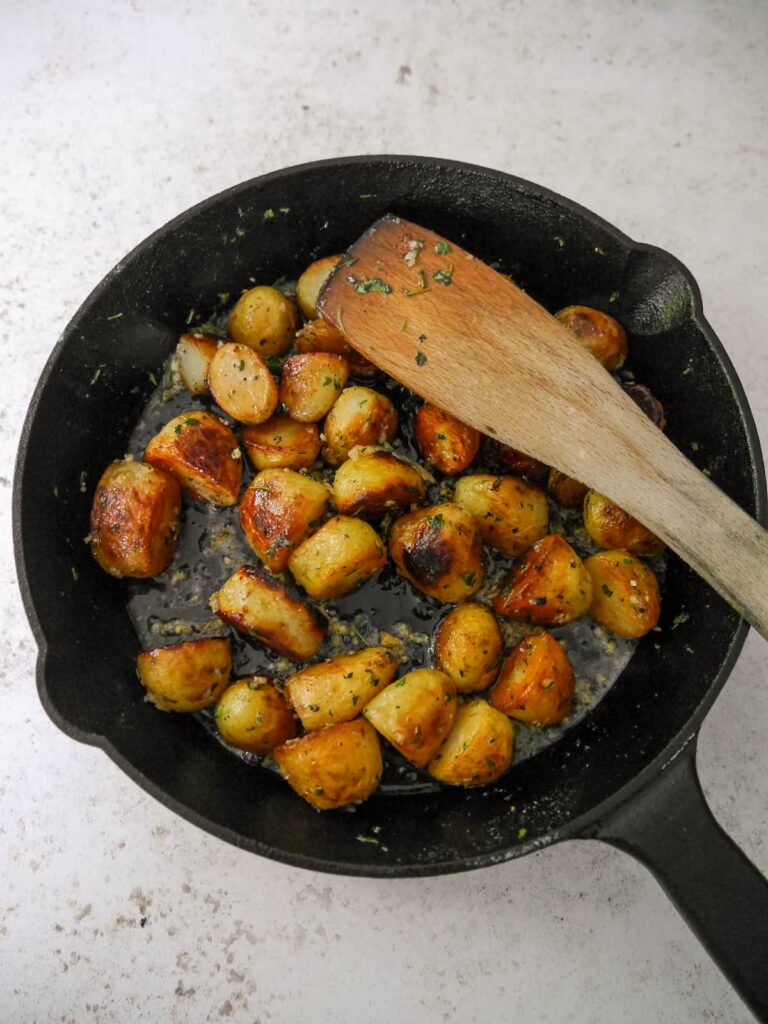 A cast iron skillet set on a wooden trivet, filled with fried potatoes in a garlic and thyme butter.