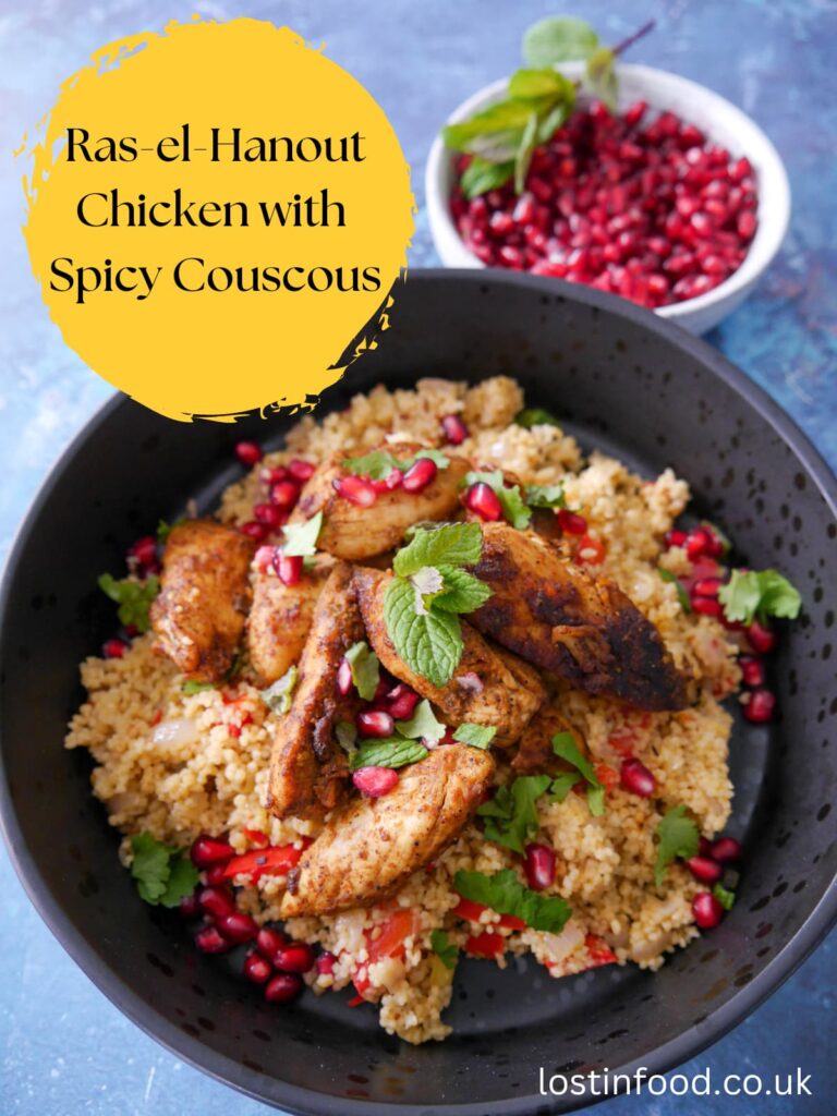 Pinnable image with recipe title and a black bowl filled with spicy couscous topped with ras-el-hanout chicken and garnished fresh herbs and pomegranate seeds, with a bowl of pomegranate seeds set alongside.