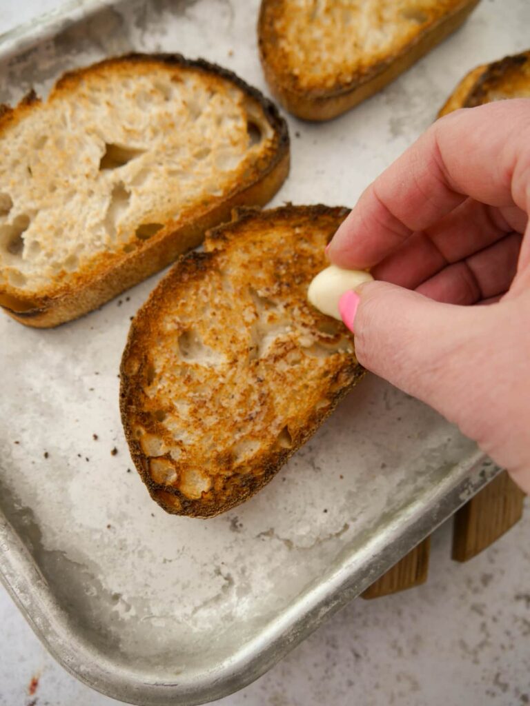 A tray of sourdough toast with raw garlic being rubbed over the toast.