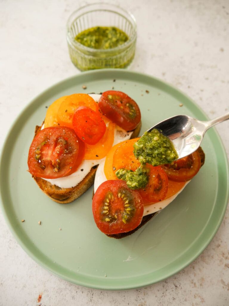 Two slices of toasted sourdough topped with whipped creme fraiche and sliced tomatoes with basil pesto being drizzled over.