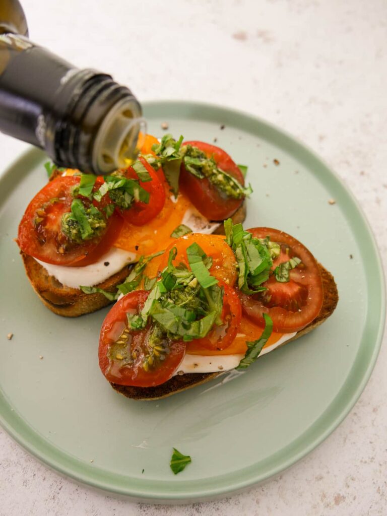 Two slices of toasted sourdough topped with whipped creme fraiche, sliced tomatoes, basil pesto and shredded basil leaves, with olive oil being drizzled over.