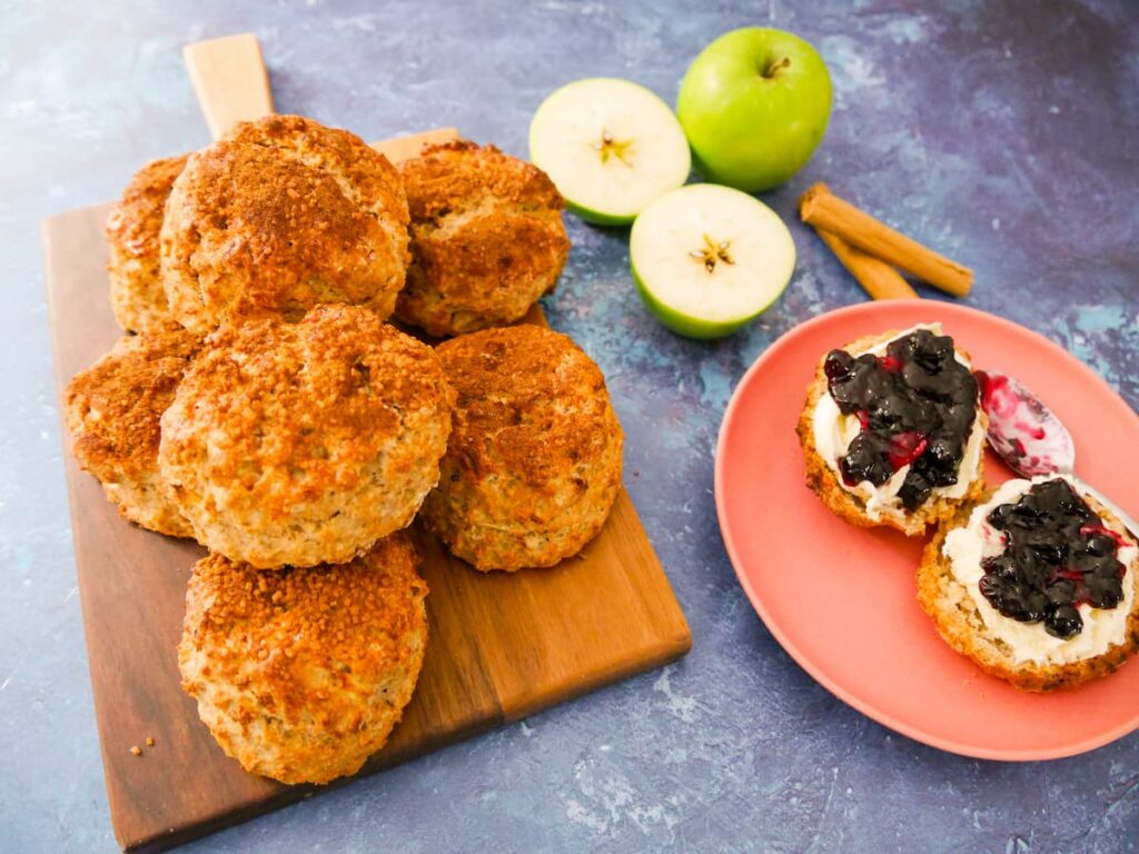 A batch of apple and cinnamon scones set on a wooden board with a pink plate with a cut scone topped with clotted cream and jam.