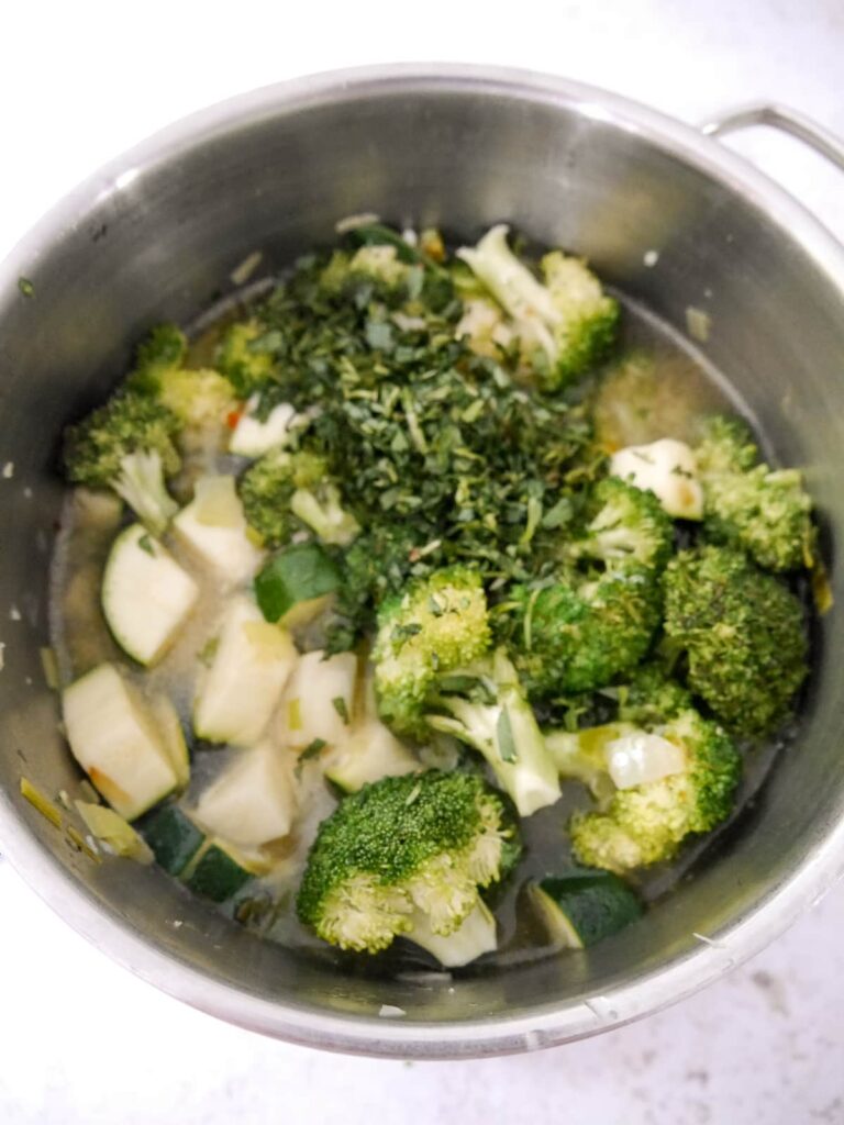 A saucepan filled with vegetable stock, broccoli florets, chunks of courgette and tarragon leaves.