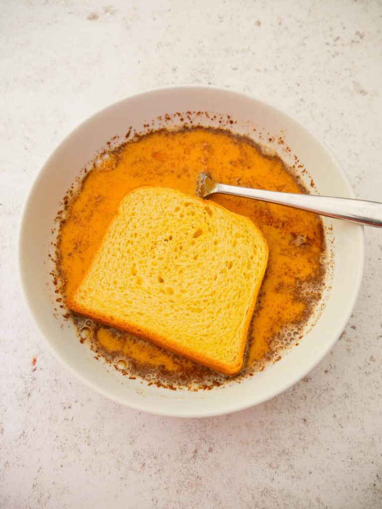 A bowl with whisked cinnamon egg mixture with a slice of brioche bread added.