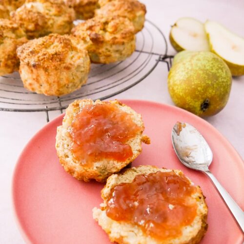 A pink plate with a pear and ginger scone topped with jam, and a wire rack of scones set alongside.