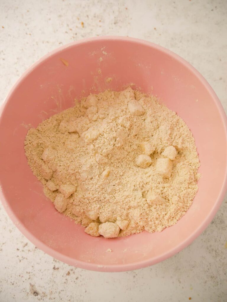 A pink mixing bowl filled with the dry ingredients for a scone, rubbed together, with added chunks of fresh pear.