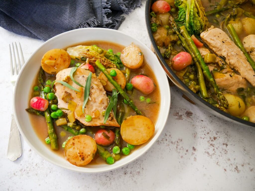 A white bowl filled with chicken and wine sauce, new potatoes, radish, asparagus and garden peas, topped with chicken breast and garnished with fresh tarragon leaves. A frying pan filled with spring chicken is set alongside.