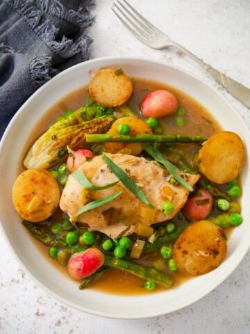 A white bowl filled with chicken and wine sauce, new potatoes, radish, asparagus and garden peas, topped with chicken breast and garnished with fresh tarragon leaves.