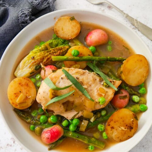 A white bowl filled with chicken and wine sauce, new potatoes, radish, asparagus and garden peas, topped with chicken breast and garnished with fresh tarragon leaves.