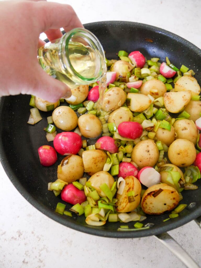 White wine being poured into a frying pan filled with sauteed potatoes, leek and garlic.