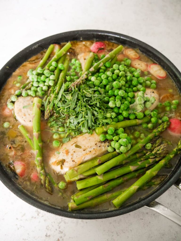 A large pan filled with sauteed chicken in a chicken stock with added radish, asparagus, garden peas and fresh tarragon leaves.