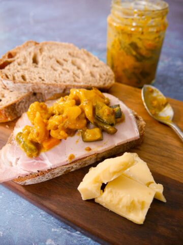 A wooden board topped with slices of sourdough bread, topped with ham and piccalilli with cheese, and a jar of piccalilli set alongside.