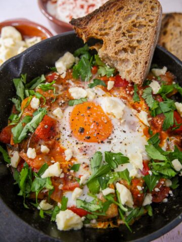 A cast iron skillet filled with mixed vegetable shakshuka topped with an egg, garnished with crumbled cheese and freshly chopped parsley, served with slices of toasted sourdough bread.